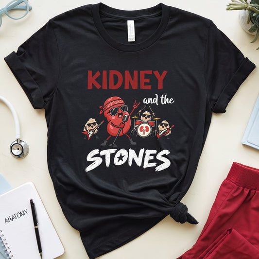 A black graphic t-shirt featuring a cartoon dog playing drums with the text 'KIDNEY and the STONES' in red and white. The shirt is styled in a flat lay with medical-themed props including a stethoscope, notebook, glasses, and red pants on a light beige background.