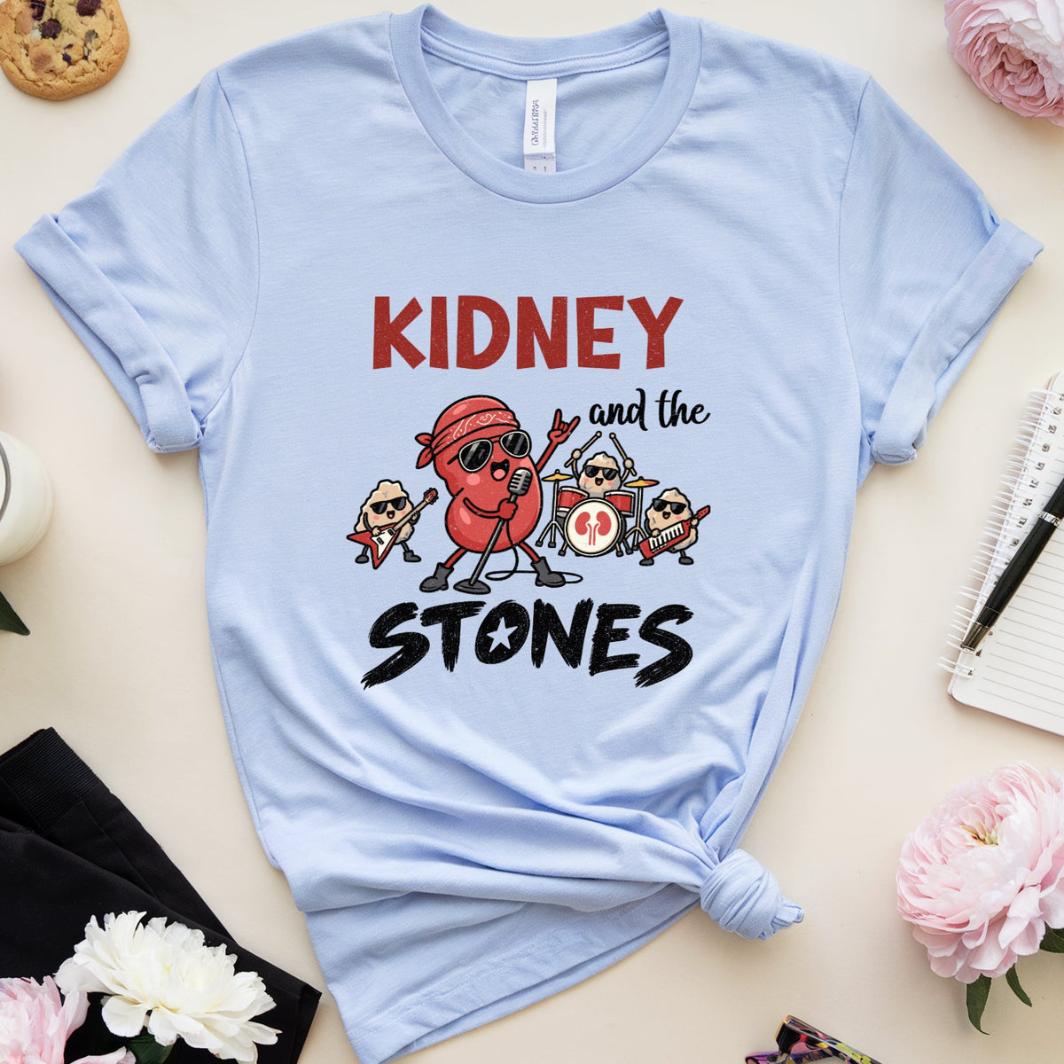 A light blue graphic t-shirt with a 'KIDNEY and the STONES' cartoon design, styled in a flat-lay arrangement with pink flowers, a cookie, a notebook, and other lifestyle props on a beige background.