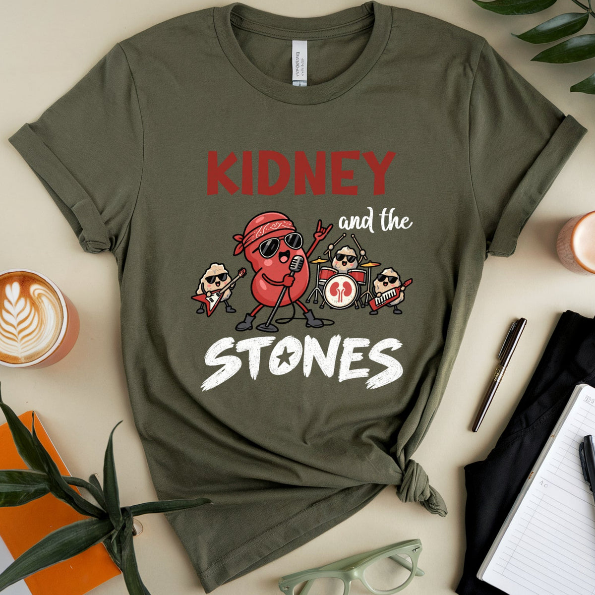 An olive green graphic t-shirt featuring a cartoon band illustration with the text 'KIDNEY and the STONES', styled in a flat lay with a coffee cup, notebook, glasses, and plants on a beige background.