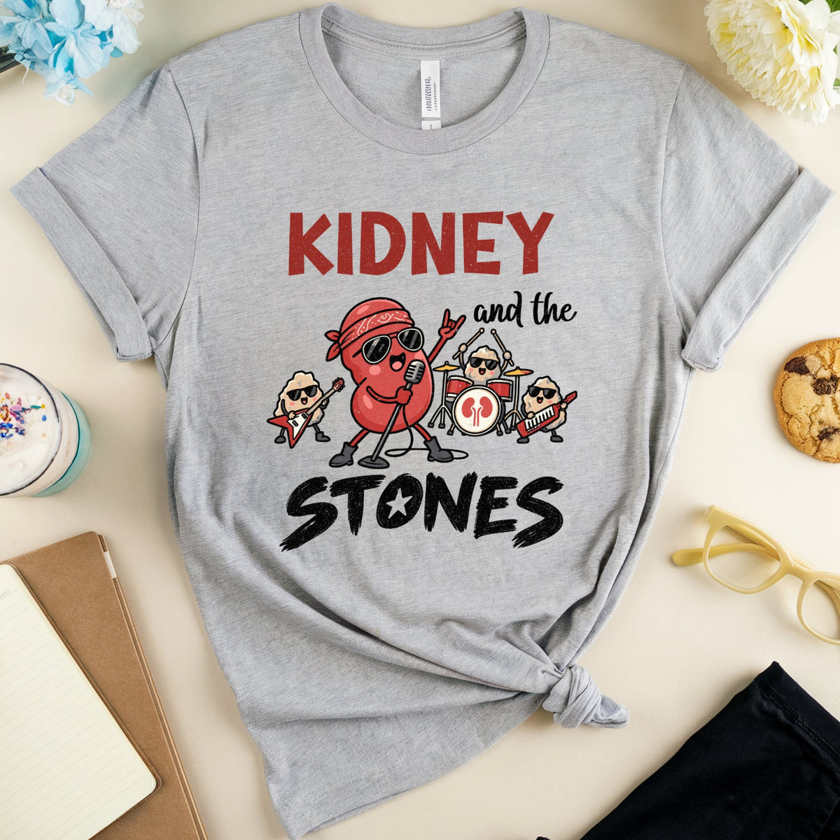 A heather gray graphic t-shirt featuring cartoon characters and the text 'KIDNEY and the STONES' in red and black. The shirt is styled in a flat-lay arrangement with a drink, notebook, flowers, sunglasses, and a cookie on a light beige background.