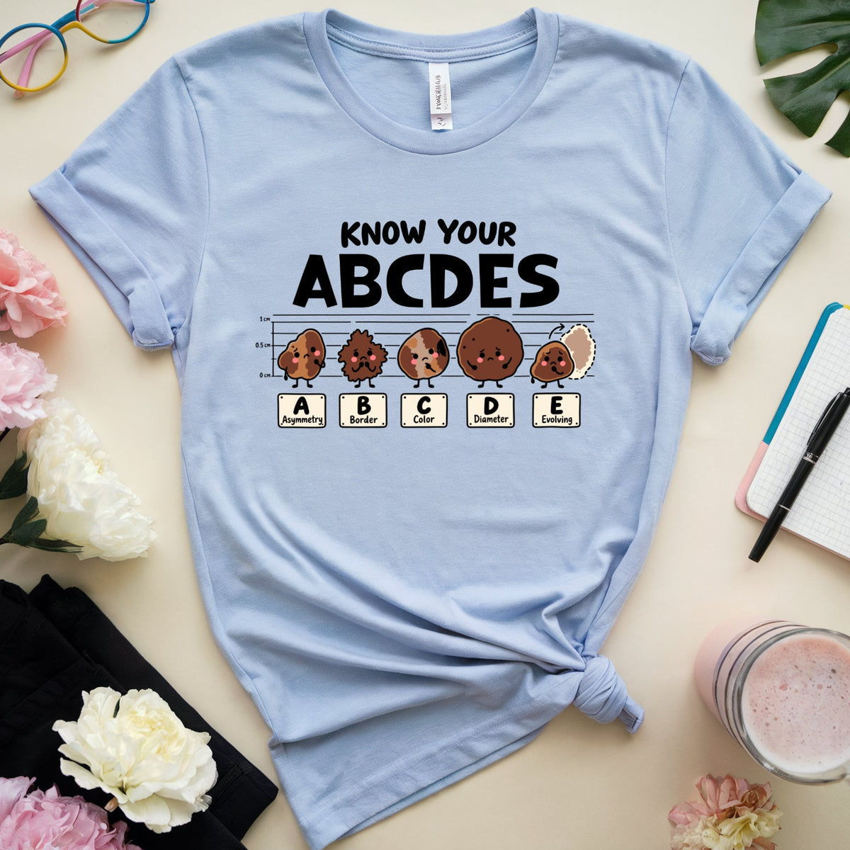 A light blue graphic t-shirt laid flat on a beige surface, surrounded by decorative props including flowers, glasses, a notebook, a drink, and a leaf. The shirt has a black text graphic reading 'KNOW YOUR ABCDES' above cartoon characters labeled A through E.