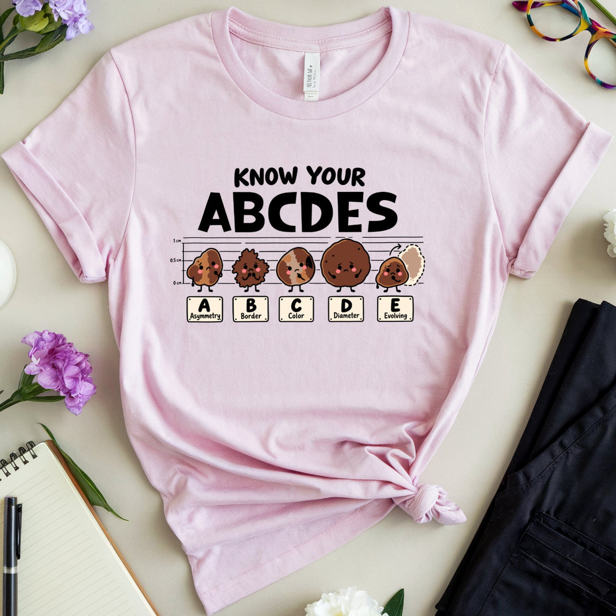 A light pink graphic t-shirt featuring a 'Know Your ABCDES' educational design, styled in a flat-lay arrangement with flowers, glasses, a notebook, and a pair of black pants on a light beige background.