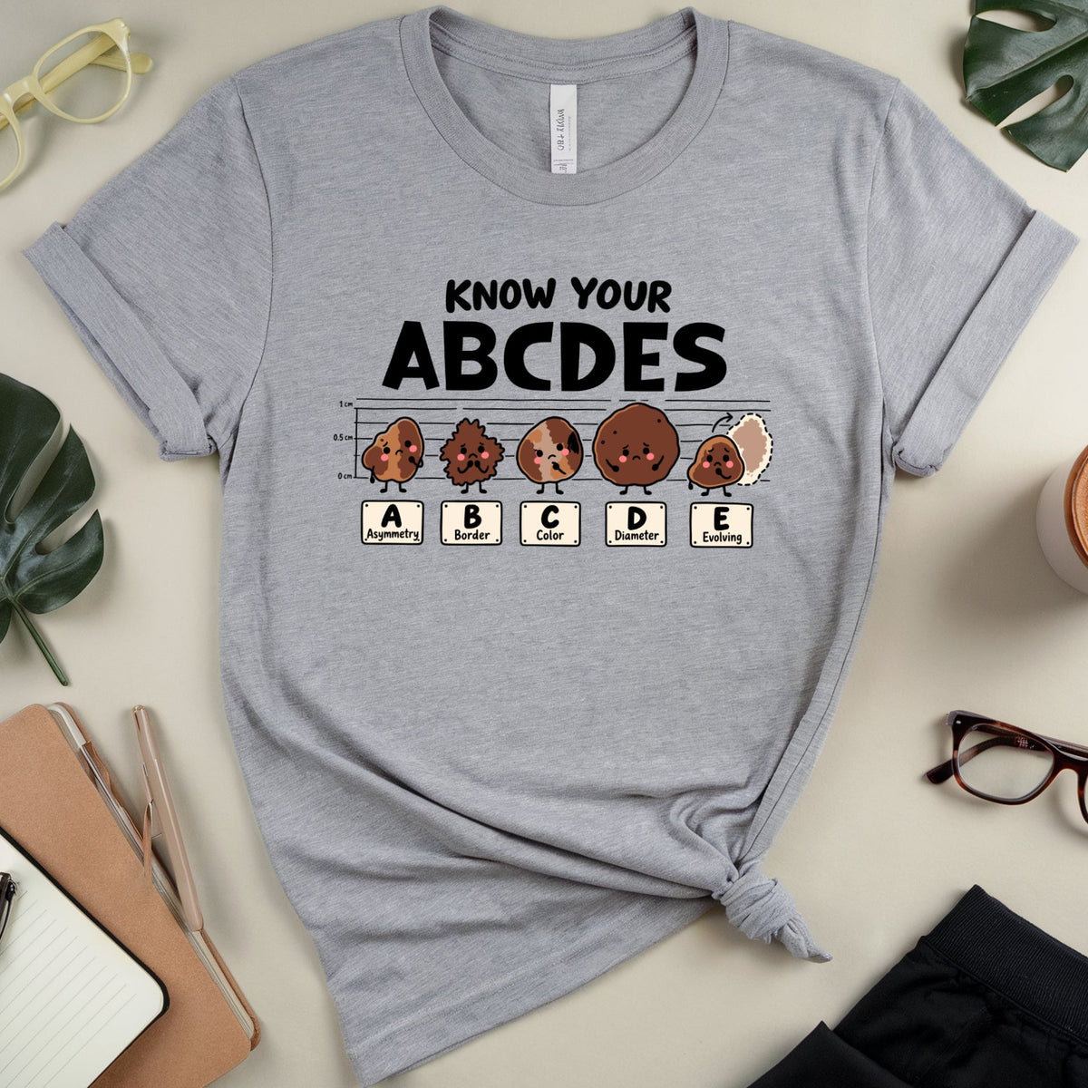 A heather gray graphic T-shirt laid flat on a light beige surface with styled props including glasses, leaves, a notebook, a cup of coffee, and folded pants. The shirt has black text reading 'KNOW YOUR ABCDES' above a row of cartoon fruit characters labeled A, B, C, D, E.
