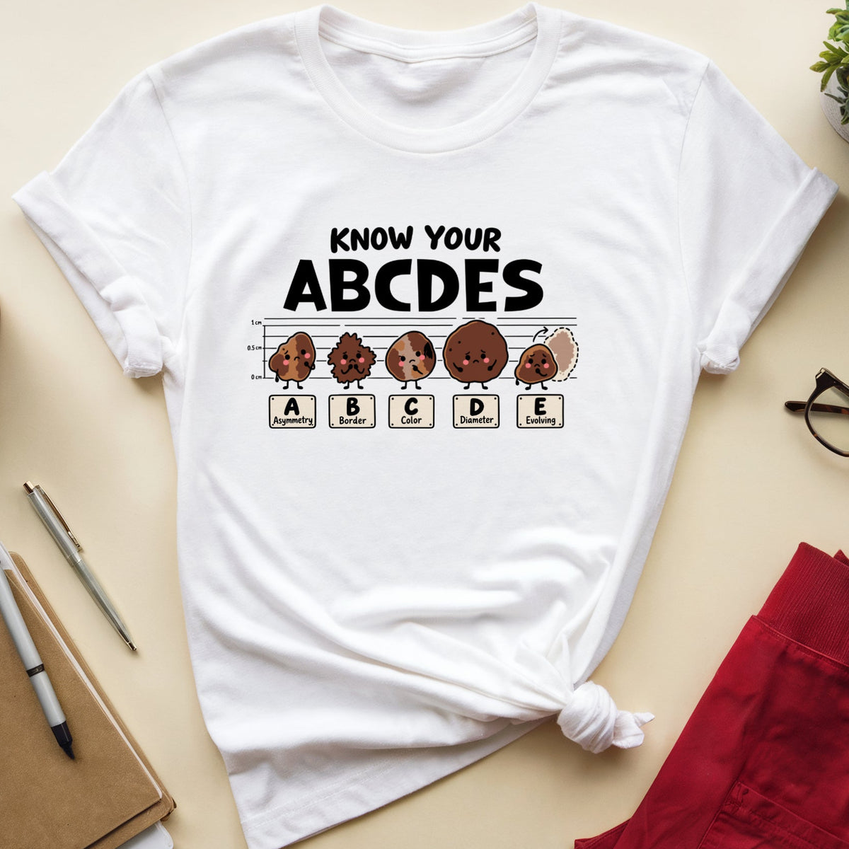 A white graphic t-shirt with a 'Know Your ABCDES' design featuring cartoon letters and musical notes, styled in a flat-lay on a beige desk with a notebook, glasses, and red pants.
