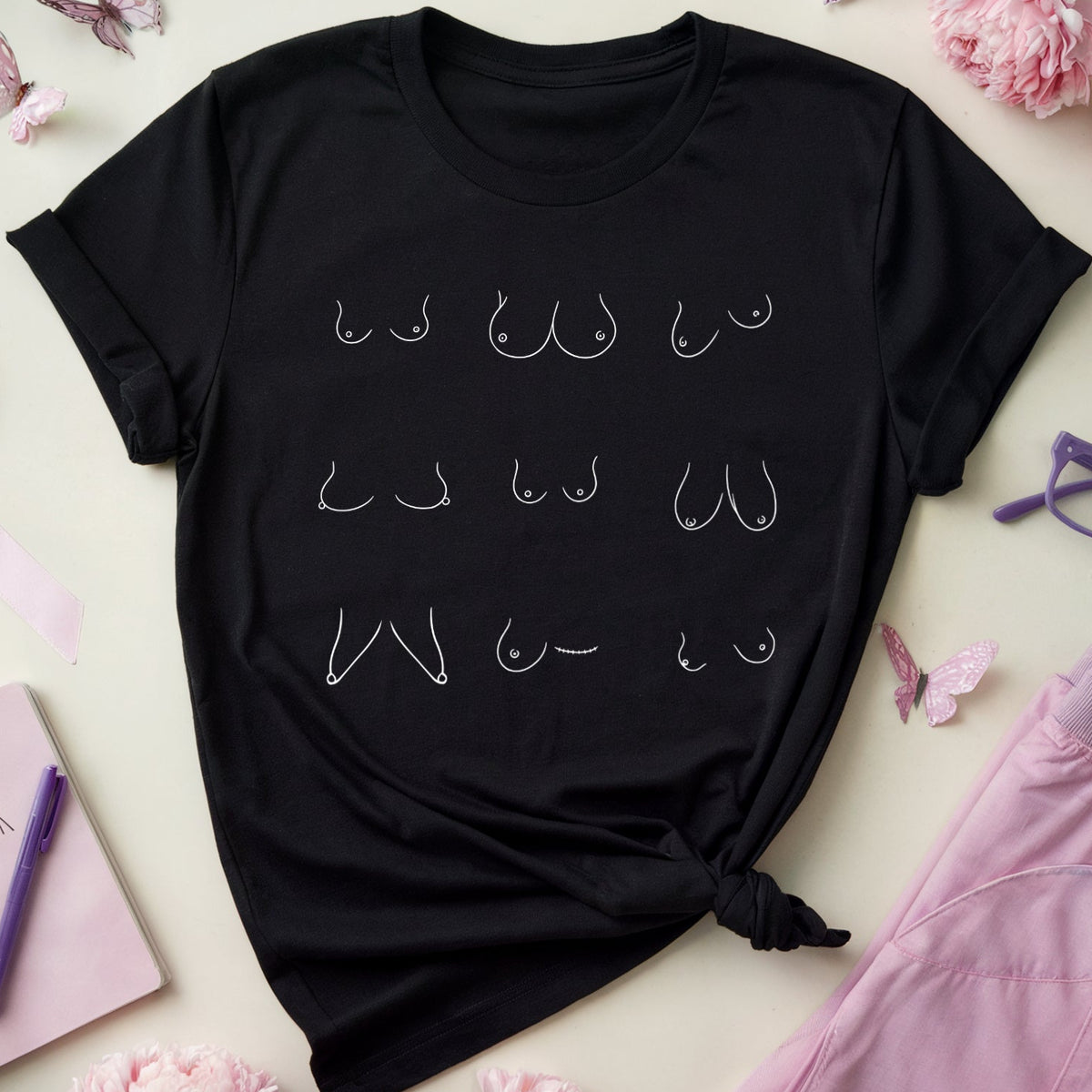 A black short-sleeve T-shirt laid flat on a light cream background, styled with pink flowers, butterflies, a pink ribbon, purple glasses, a pink notebook, and lavender scrubs. The shirt has a white graphic of repeated female nipple shapes across the chest.