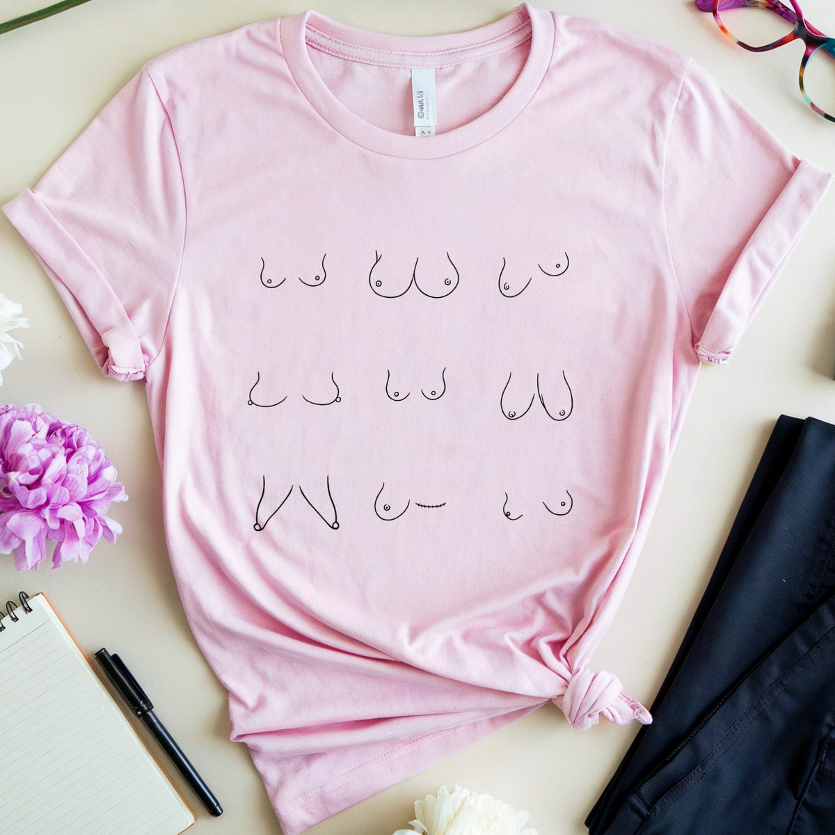 A light pink short-sleeve t-shirt with a graphic of repeated bare nipple illustrations, styled in a flat lay with flowers, eyeglasses, a notebook, a pen, a tube of lipstick, and a pair of dark pants on a beige background.