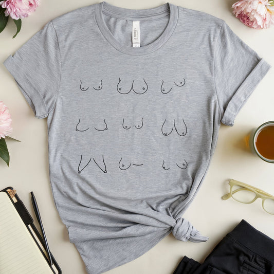 A heather gray graphic t-shirt with a knotted hem, featuring a black line art print of various bra styles. The t-shirt is styled in a flat-lay arrangement with pink flowers, a notebook, glasses, a cup of tea, and black pants on a light beige background.