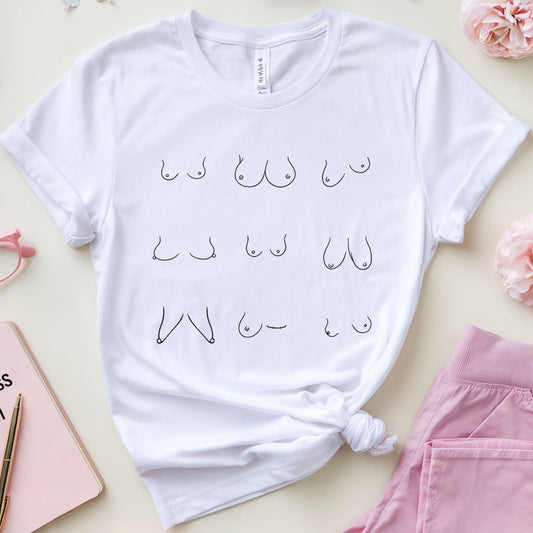 A styled flat-lay of a white graphic T-shirt with black Arabic calligraphy, surrounded by pink and white flowers, pink glasses, a pink awareness notebook, and folded pink pants on a light beige background.