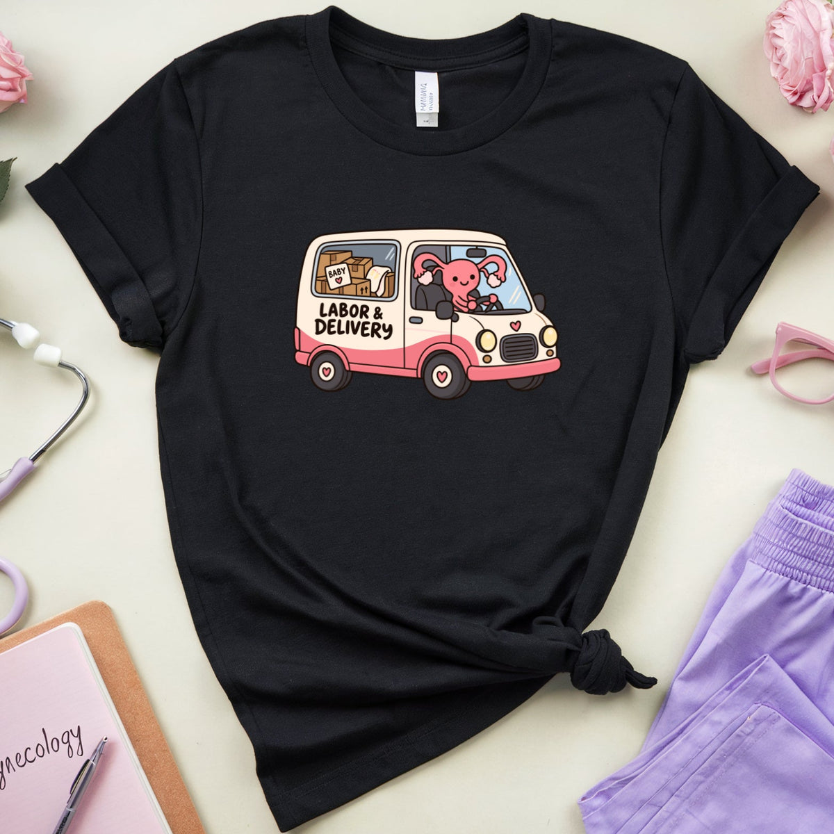 A black graphic t-shirt featuring a cute pink and white van with cartoon animals and the text 'LABOR & DELIVERY'. The shirt is styled in a flat-lay arrangement with lavender scrubs pants, pink glasses, a purple stethoscope, a pink notebook, and pink roses on a light beige background.