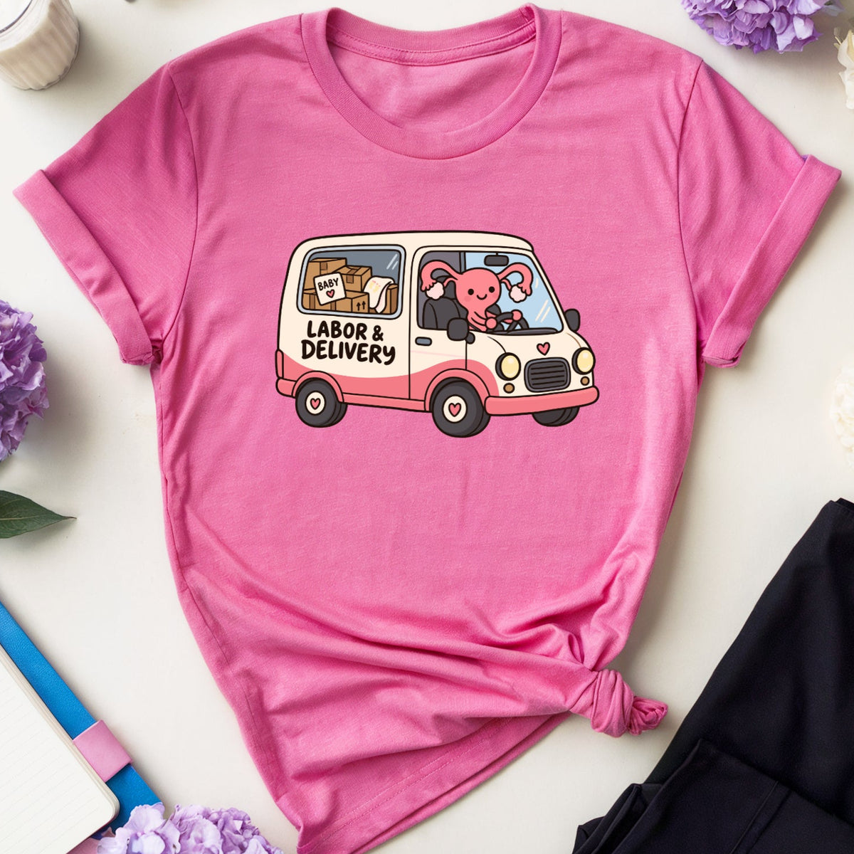 A pink short-sleeve T-shirt laid flat on a light surface, styled with flowers, glasses, a drink, a notebook, and black pants. The shirt has a cartoon graphic of a pink bear driving a cream van with the words 'LADE & DELIVRS'.