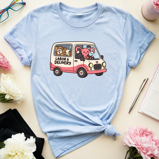A light blue graphic t-shirt with a cute cartoon van design, styled in a flat-lay arrangement with flowers, glasses, a notebook, and a pen on a cream-colored background.