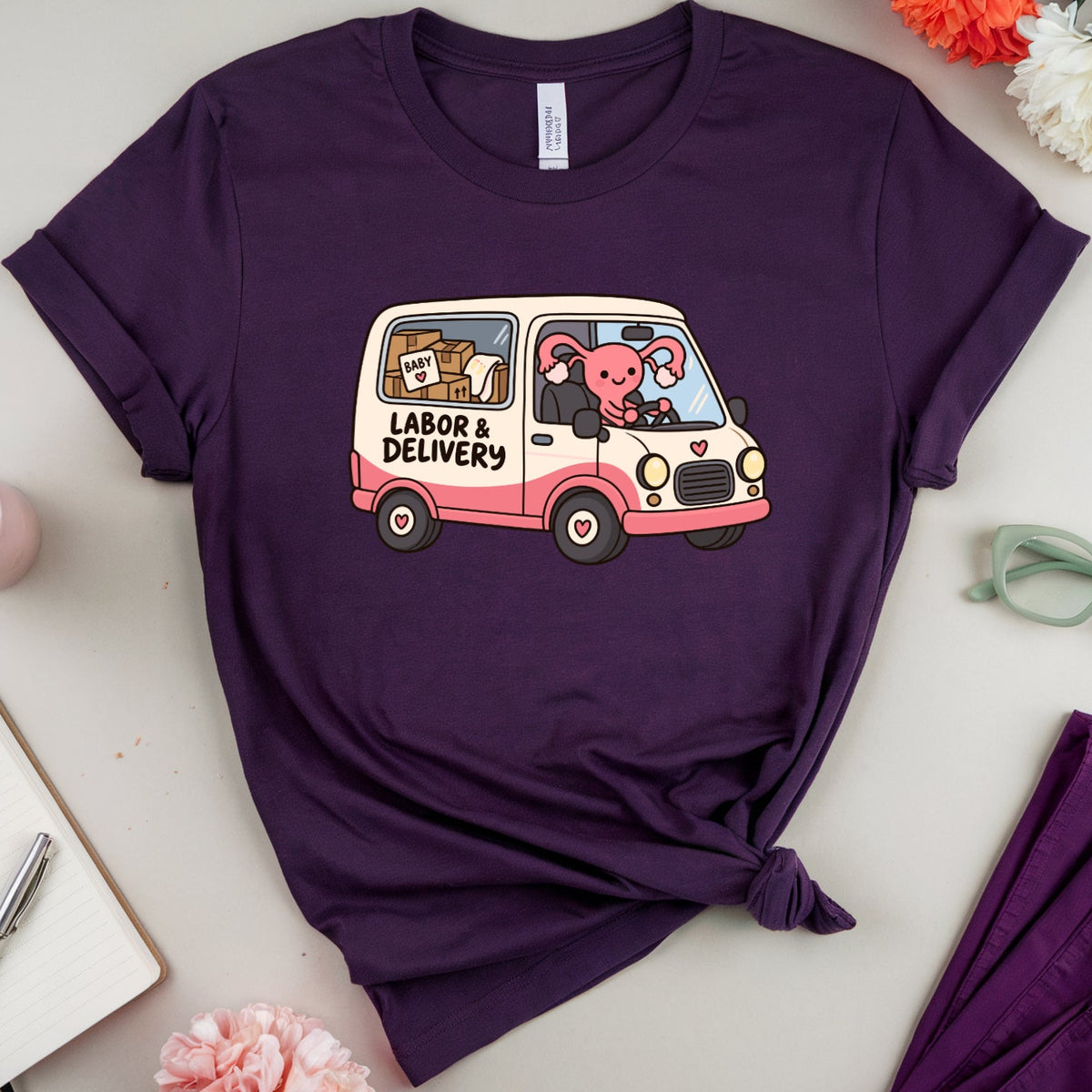 A purple short-sleeve crewneck t-shirt with a graphic of a pink and white 'Labor & Delivery' van carrying cartoon animals, styled in a flat-lay arrangement with flowers, glasses, a notebook, and a matching purple garment.
