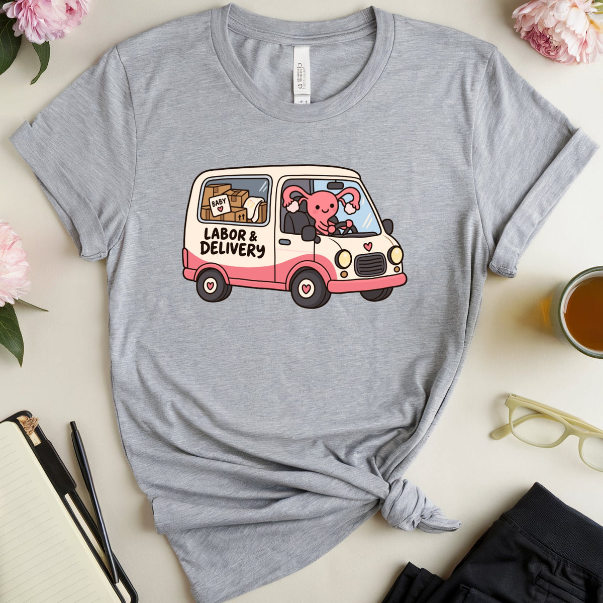 A heather gray graphic T-shirt laid flat in a styled flat-lay arrangement. The shirt has a cartoon van graphic with the words 'LABOR & DELIVERY' and a pink pig driving.