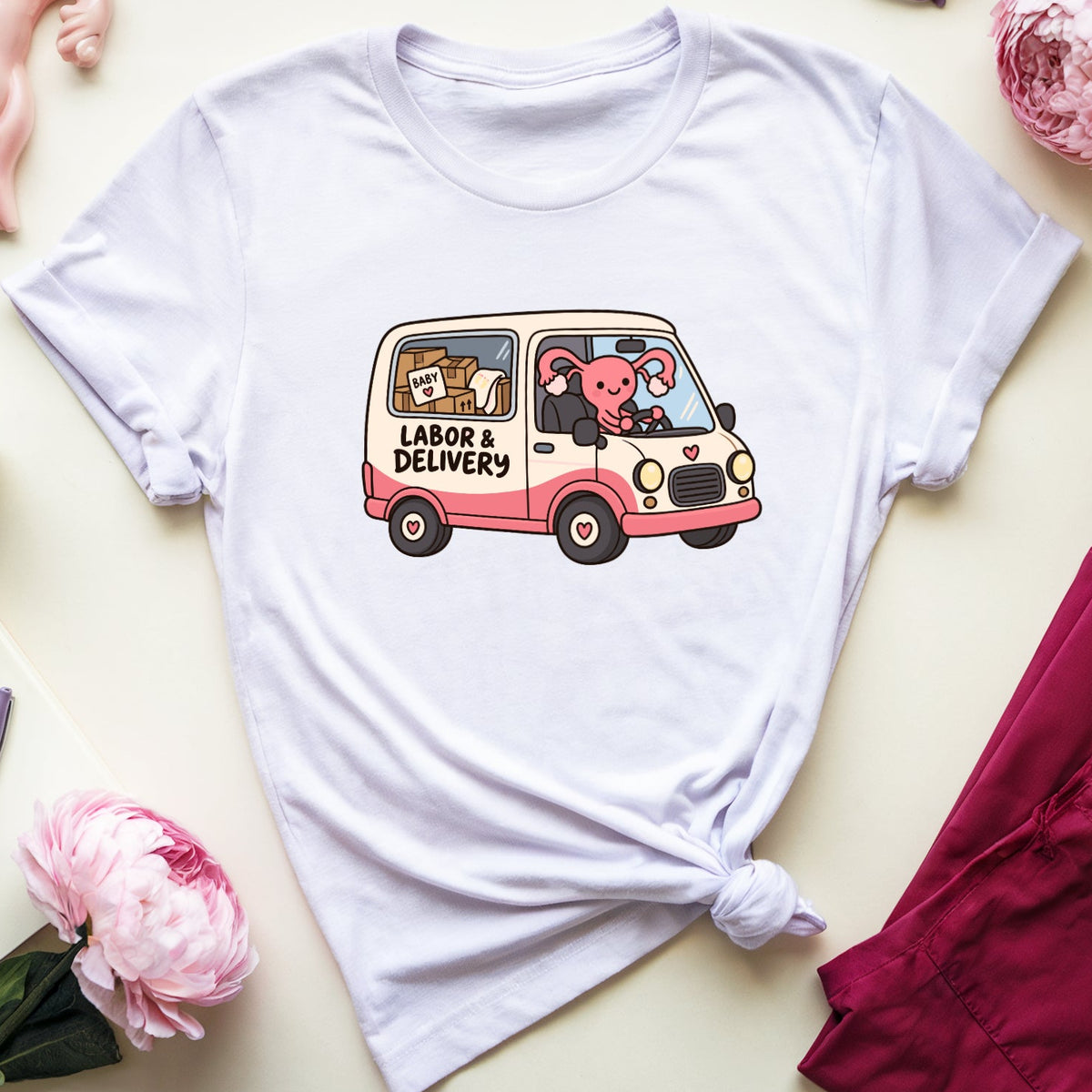 A light lavender graphic t-shirt featuring a cartoon illustration of a pink and white van with the words 'LABOR & DELIVERY' and cute animal characters. The t-shirt is styled in a flat lay with pink and white peonies, a pink anatomical model, purple eyeglasses, a notebook, and a pair of burgundy pants on a warm cream background.