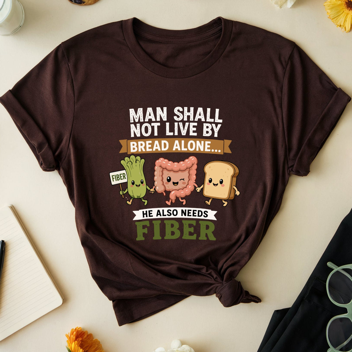 A dark brown graphic t-shirt is displayed in a flat-lay arrangement on a light beige background. The shirt features a humorous design with cartoon bacteria and bread slices, accompanied by the text 'MAN SHALL NOT LIVE BY BREAD ALONE.
