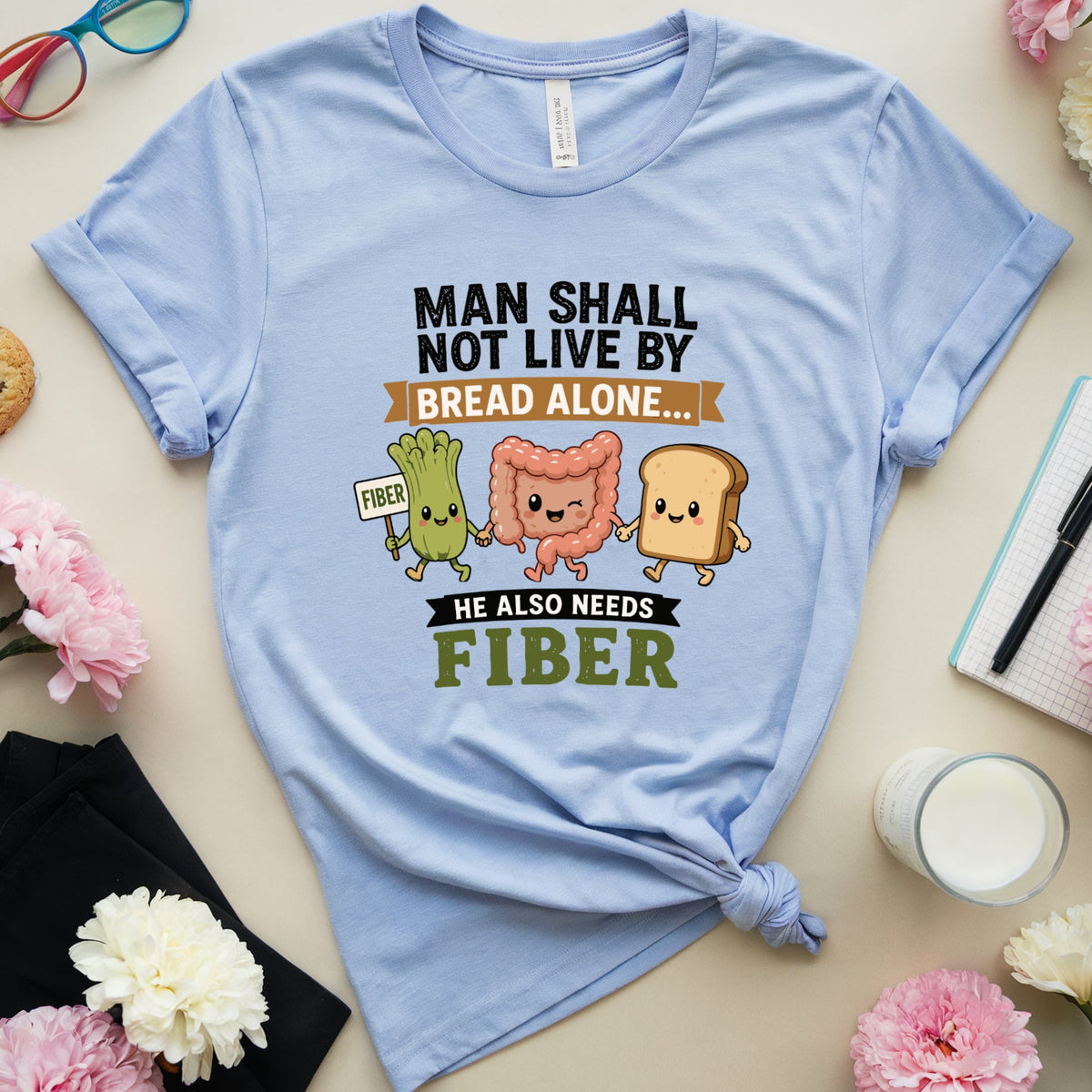 A light blue graphic t-shirt with a 'Fiber' theme, styled in a flat-lay arrangement with flowers, glasses, a notebook, and a cookie on a beige background.