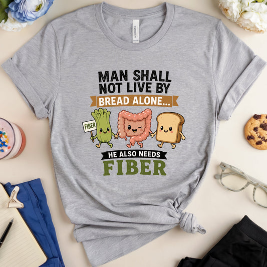 A heather gray graphic T-shirt laid flat on a light beige surface, styled with a drink, notebook, glasses, cookie, and flowers around it. The shirt has a printed slogan reading “MAN SHALL NOT LIVE BY BREAD ALONE.