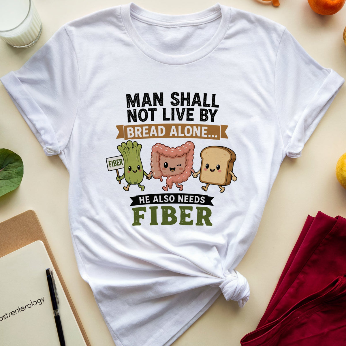 A white graphic T-shirt laid flat on a light beige surface, surrounded by props including a notebook, glasses, fruit, leaves, and a folded red garment. The shirt has a printed slogan reading “MAN SHALL NOT LIVE BY BREAD ALONE.
