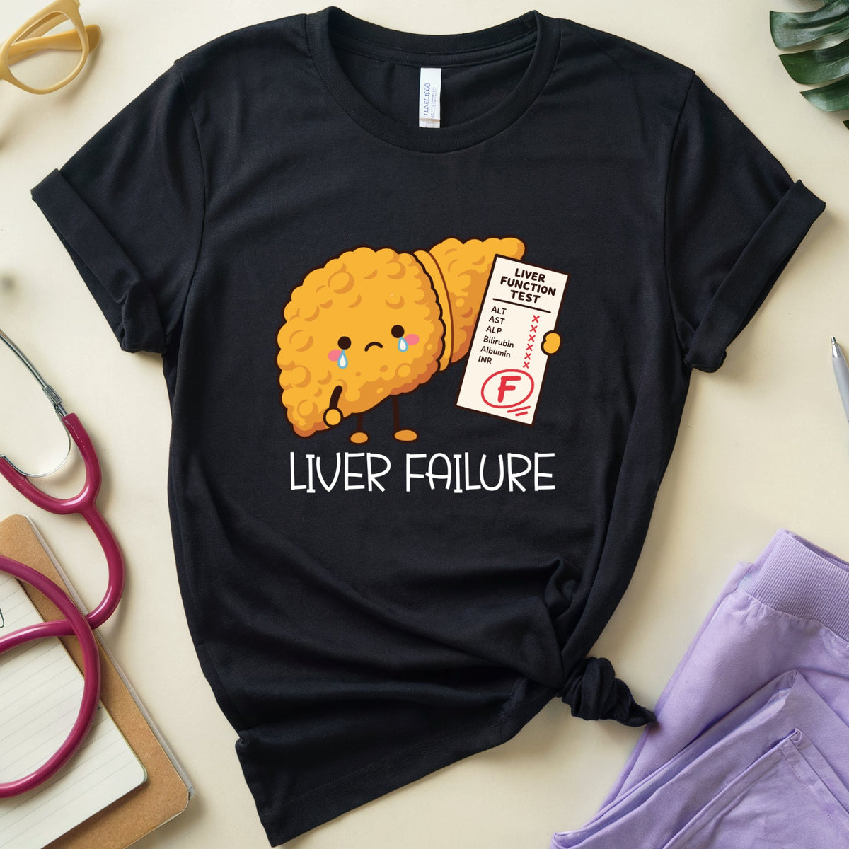 A black short-sleeve graphic T-shirt laid flat on a light beige surface, styled with medical-themed props including a stethoscope, glasses, a pen, a notebook, and folded lavender scrubs. The shirt features a cartoon liver graphic holding an F-grade report card, with the text 'LIVER FAILURE' below.