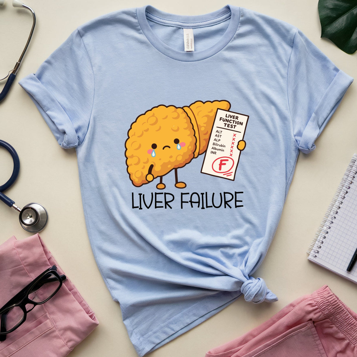 A light blue graphic t-shirt featuring a cute cartoon liver character with a failing G test sheet, surrounded by medical-themed flat-lay props including a stethoscope, pink pants, glasses, and a notebook on a beige background.