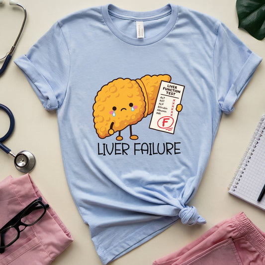 A light blue graphic t-shirt featuring a cute cartoon liver character with a failing G test sheet, surrounded by medical-themed flat-lay props including a stethoscope, pink pants, glasses, and a notebook on a beige background.