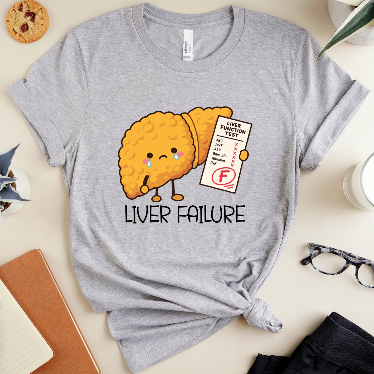 A heather gray graphic t-shirt featuring a cute crying liver character holding an F-grade report card, with the text 'LIVER FAILURE' underneath. The t-shirt is styled in a flat-lay arrangement with a notebook, glasses, a plant, and other desk accessories on a light beige background.