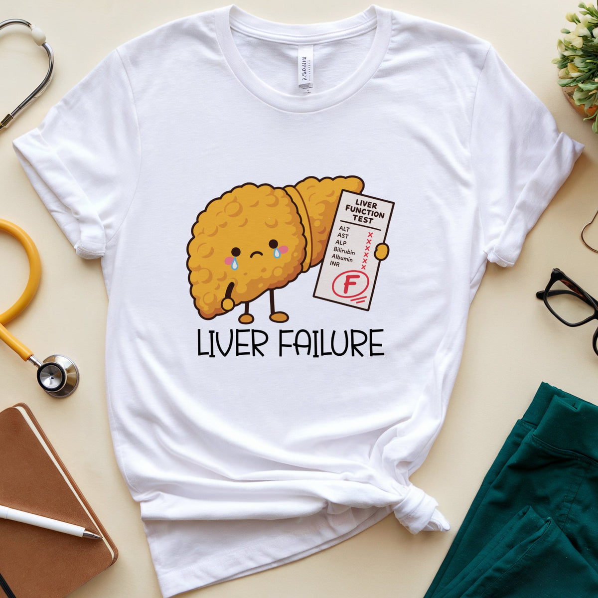 A white graphic t-shirt with a cartoon liver character holding a failing probation test, styled in a flat lay with medical props like a stethoscope, notebook, glasses, and scrub pants on a beige background.