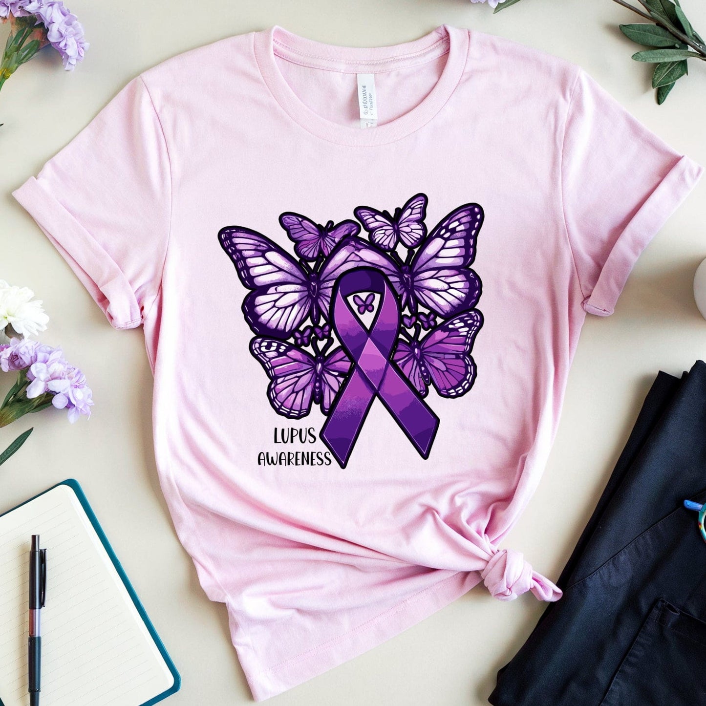 Pink t-shirt with purple butterfly and ribbon design on a light surface with flowers and a cup.