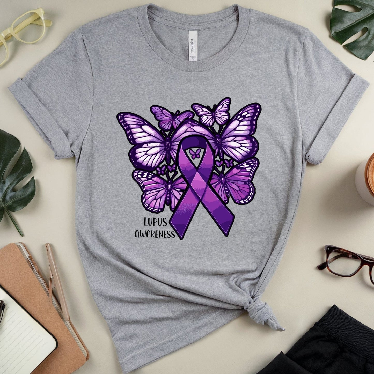 Gray t-shirt with purple butterfly and awareness ribbon design on a light surface with accessories.