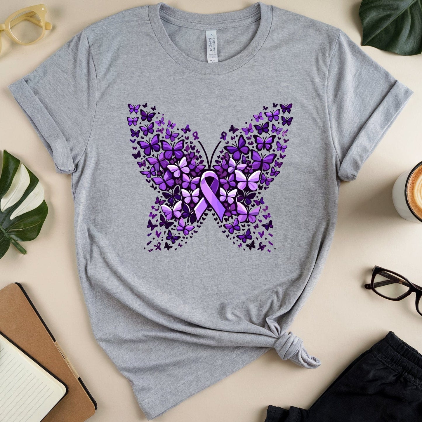 Gray t-shirt with purple butterfly design on a beige surface with accessories.