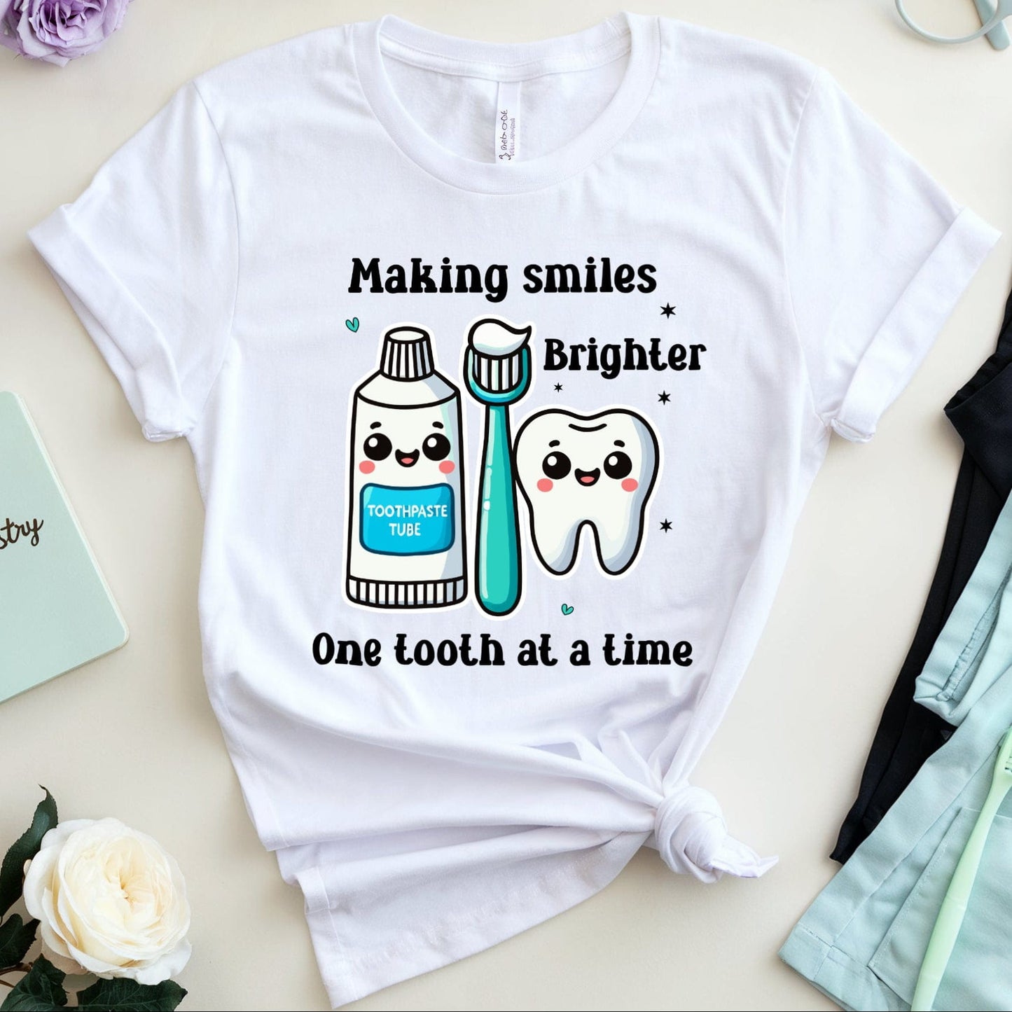 White t-shirt with dental-themed design and text on a light surface with flowers and glasses.
