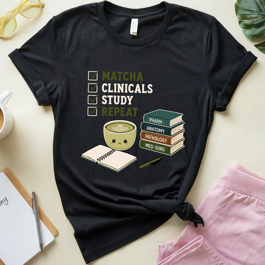 A black graphic t-shirt laid flat on a light beige surface, styled with a coffee cup, yellow glasses, a notebook, pink shorts, and a green leaf. The t-shirt features a checklist graphic reading 'MATCHA CLINICALS STUDY REPEAT' next to a coffee cup, a smiling notebook, and a stack of books.