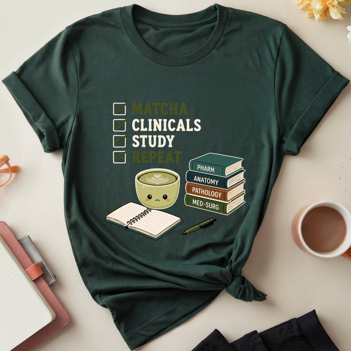 A dark green graphic t-shirt styled in a flat lay with a checklist design reading 'MATCHA CLINICALS STUDY REPEAT' alongside a coffee cup, notebook, books, glasses, and flowers on a light beige background.