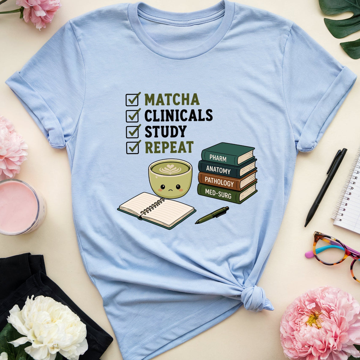 A light blue graphic t-shirt is displayed in a flat-lay arrangement on a beige surface. The shirt features a cute cartoon illustration of a matcha bowl next to a stack of books, with a checklist reading 'MATCHA CLINICALS STUDY REPEAT'.