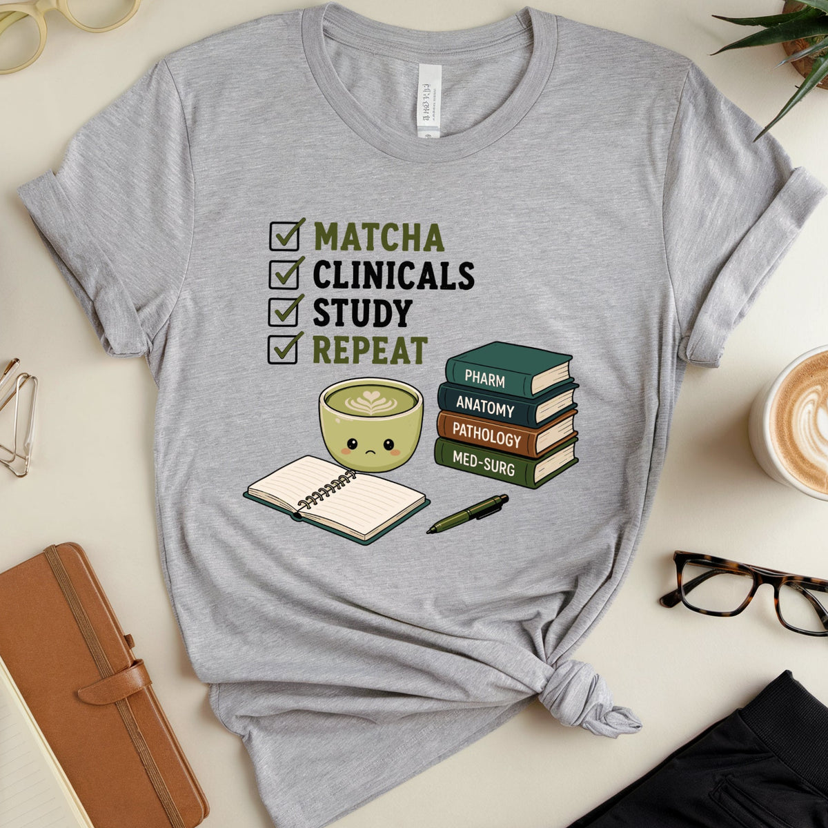 A heather gray graphic t-shirt is displayed in a styled flat-lay arrangement on a light beige surface. The shirt features a graphic of a green matcha bowl, study books, and a checklist reading 'MATCHA CLINICALS STUDY REPEAT'.