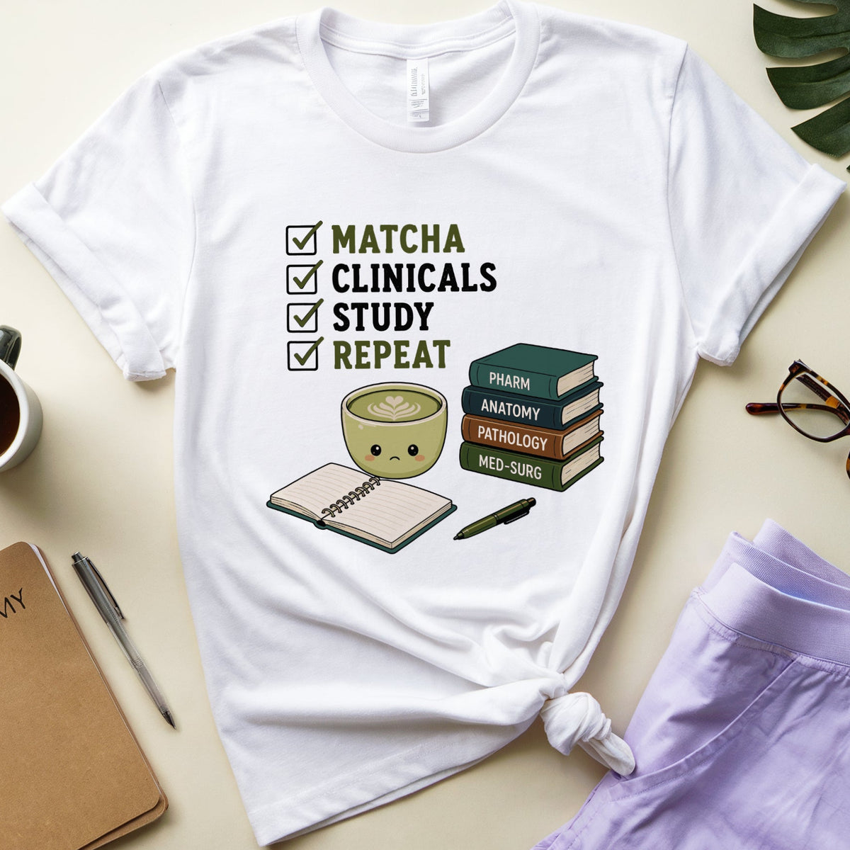 A white graphic t-shirt featuring a medical school study checklist design with a coffee cup, books, and notebook illustration. The t-shirt is styled in a flat-lay arrangement with lavender shorts, a notebook, eyeglasses, a cup of coffee, and a green leaf on a light beige background.