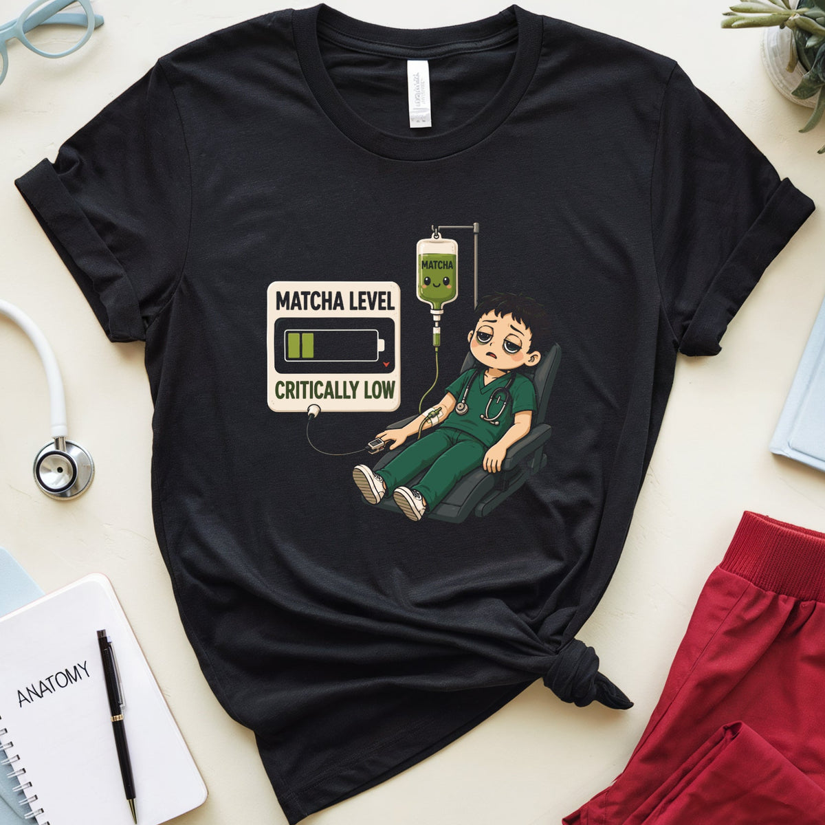 A black graphic t-shirt featuring a cartoon illustration of a nurse with an IV drip and the text 'MATCHA LEVEL CRITICALLY LOW'. The t-shirt is styled in a flat lay on a light beige surface with medical-themed props including a stethoscope, notebook, glasses, and red scrubs.