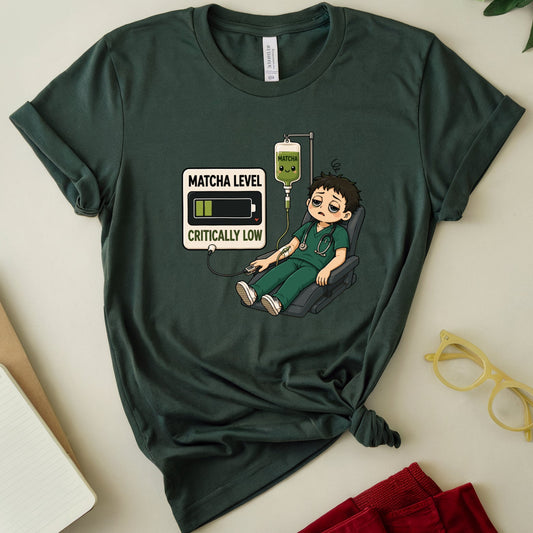 A dark green graphic t-shirt featuring a cartoon illustration of a doctor with a battery icon and the text 'MATCHA LEVEL CRITICALLY LOW'. The t-shirt is styled in a flat-lay arrangement on a light beige background with a notebook, glasses, and red pants.