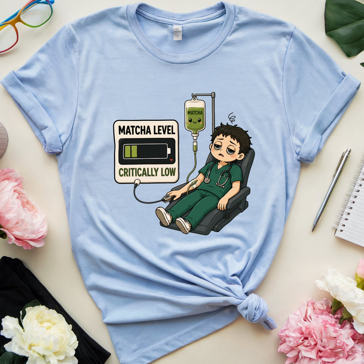A light blue graphic t-shirt featuring a cartoon illustration of a nurse with a battery icon for matcha, surrounded by pink and white flowers, glasses, and a notebook in a flat-lay arrangement.