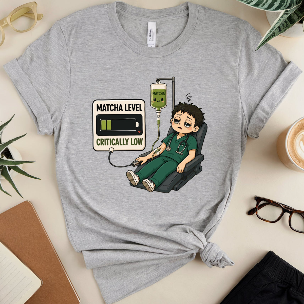 A heather gray graphic T-shirt laid flat on a light beige surface, styled with glasses, plants, a notebook, a cup of coffee, and black pants. The shirt has a cartoon graphic of a tired nurse sitting in a chair with an IV bag and a battery icon, with text reading 'MATCHA LEVEL CRITICALLY LOW'.