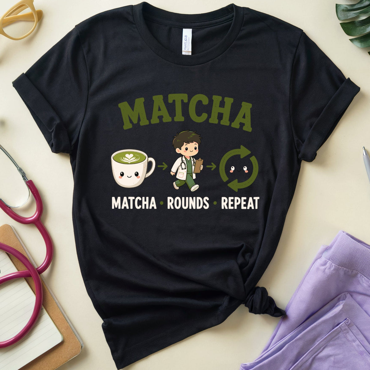 A black graphic t-shirt featuring a cute matcha-themed cartoon design with the text 'MATCHA MATCHA ROUNDS REPEAT'. The shirt is styled in a flat-lay arrangement on a beige surface with medical and lifestyle props including a stethoscope, glasses, a plant leaf, and purple scrubs.