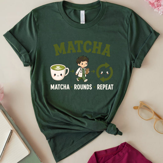 A dark green graphic t-shirt featuring a cute cartoon matcha-themed design with the text 'MATCHA MATCHA ROUNDS REPEAT' and illustrations of a matcha cup and a matcha worker. The t-shirt is styled in a flat-lay arrangement on a light beige background with pink flowers, a notebook, glasses, and a magenta garment.