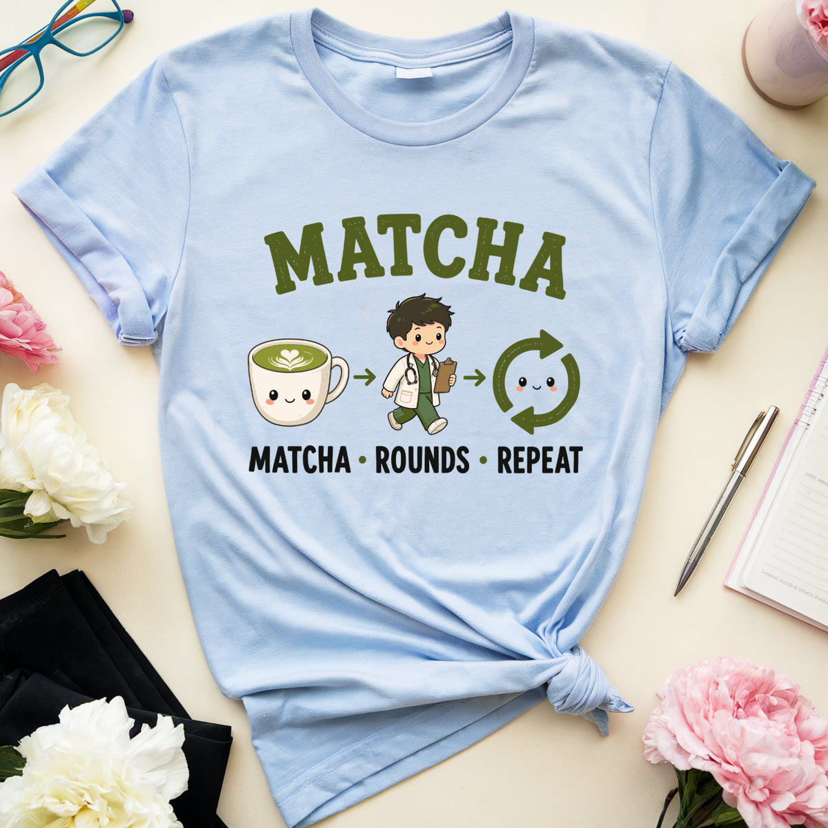 A light blue graphic t-shirt with a 'MATCHA' theme, styled in a flat-lay arrangement with pink and white peonies, eyeglasses, a notebook, and a pen on a cream-colored background.
