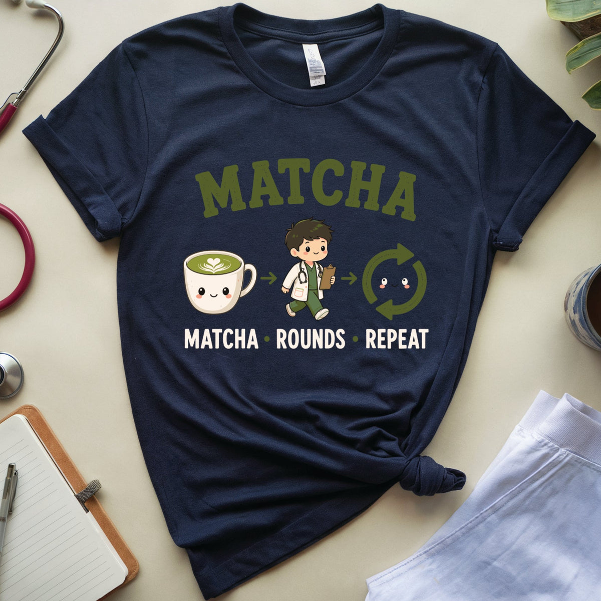A navy blue graphic t-shirt featuring a 'MATCHA' design with a cute cartoon doctor, a smiling matcha cup, and the words 'MATCHA ROUNDS REPEAT'. The shirt is styled in a flat-lay arrangement with a stethoscope, notebook, plant, and coffee cup on a light beige background.