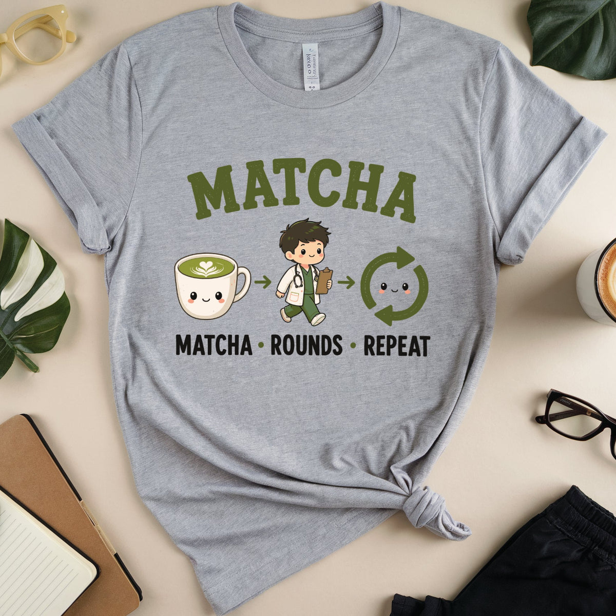 A heather gray graphic t-shirt featuring a cute cartoon matcha-themed design with the text 'MATCHA MATCHA ROUNDS REPEAT'. The shirt is styled in a flat-lay arrangement with a coffee cup, glasses, notebook, and leaves on a beige background.