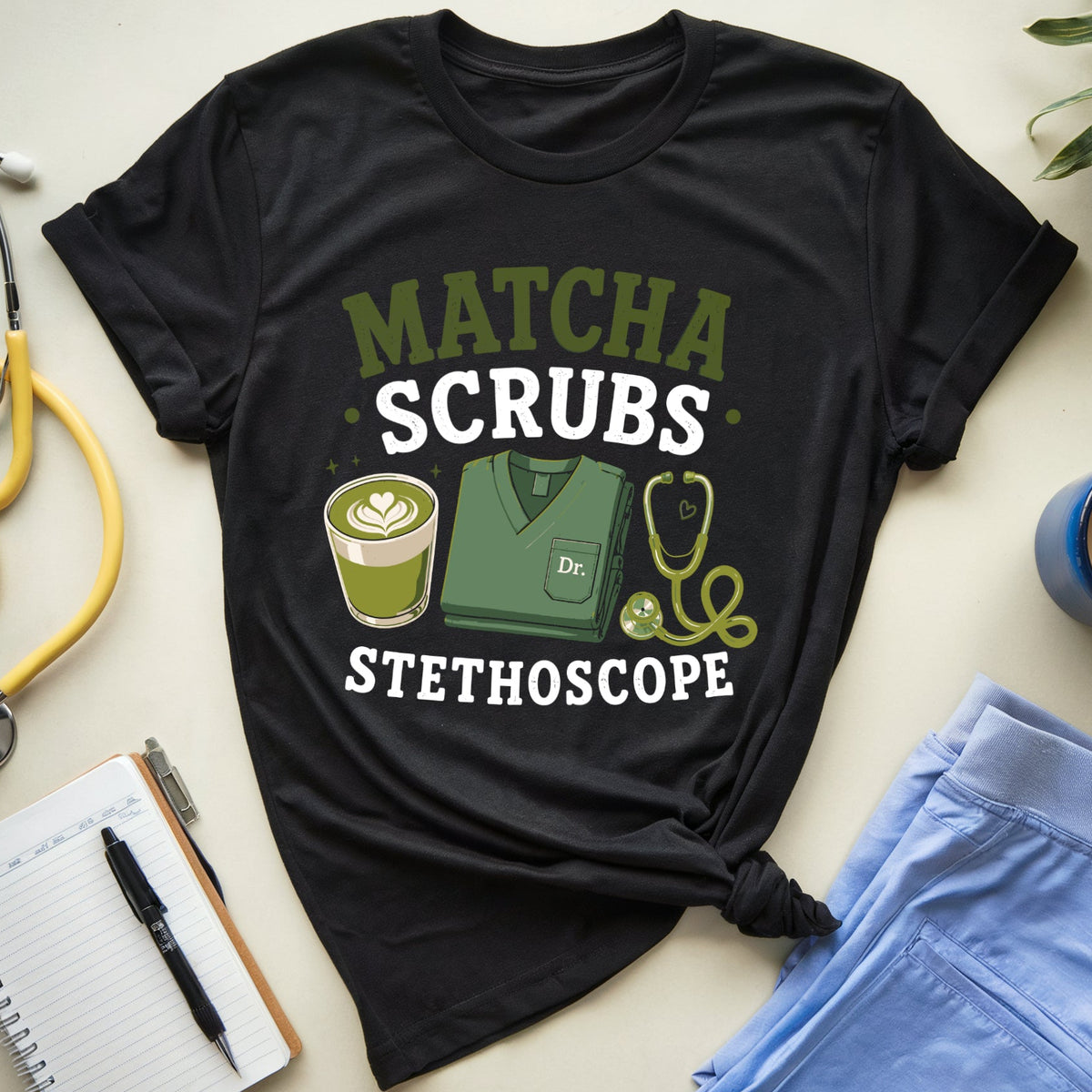 A black graphic T-shirt laid flat on a light beige surface, surrounded by medical-themed props including a yellow stethoscope, a notebook, pen, blue scrubs, a coffee mug, and a plant. The shirt reads “MATCHA SCRUBS STETHOSCOPE” with a matcha latte, stethoscope, and scrub top illustration.