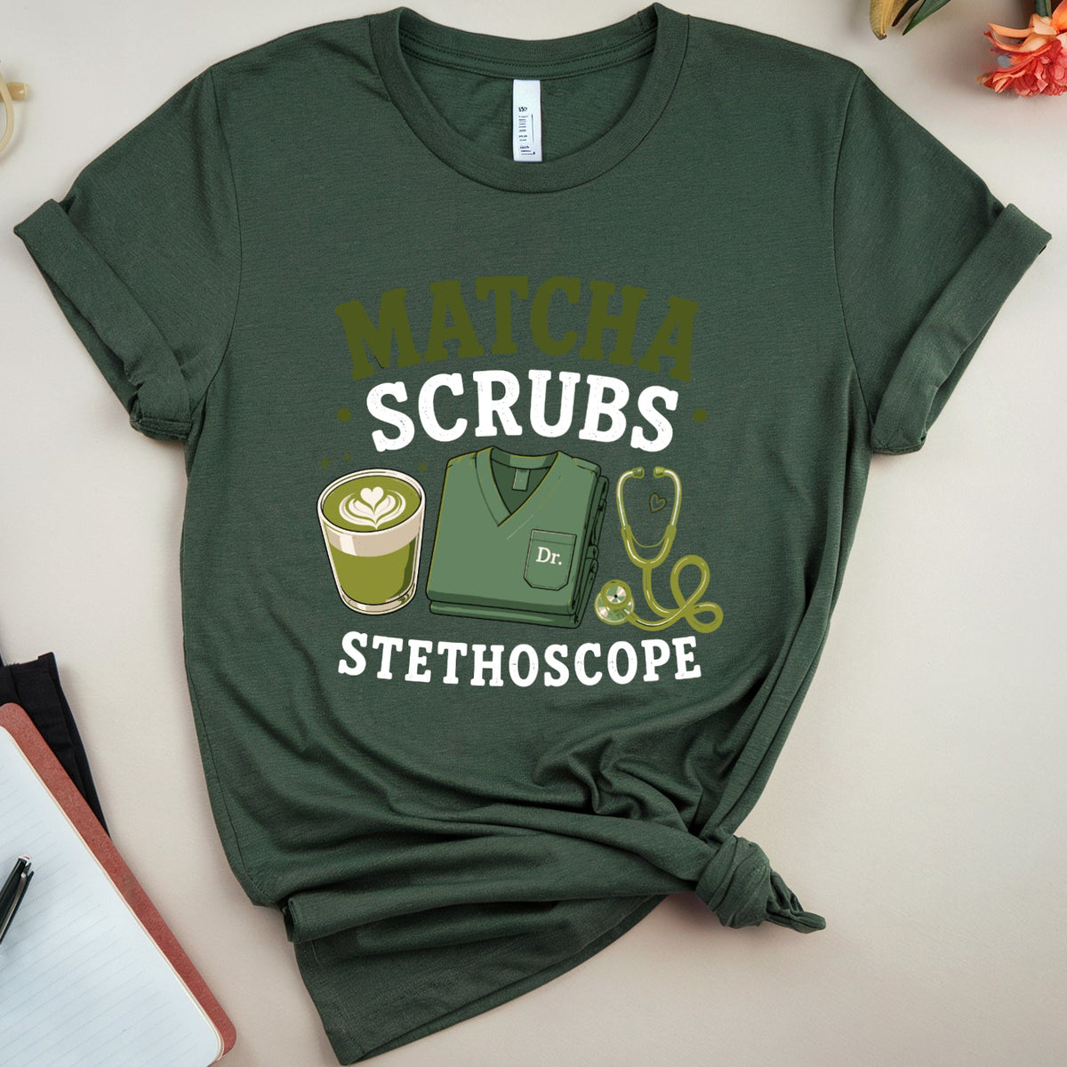 A dark green graphic t-shirt styled in a flat lay with a knotted hem, featuring the words 'MATCHA SCRUBS' and 'STETHOSCOPE' with illustrations of a coffee cup, stethoscope, and scrub top. The shirt is displayed on a light beige background with decorative props including glasses, a notebook, and flowers.
