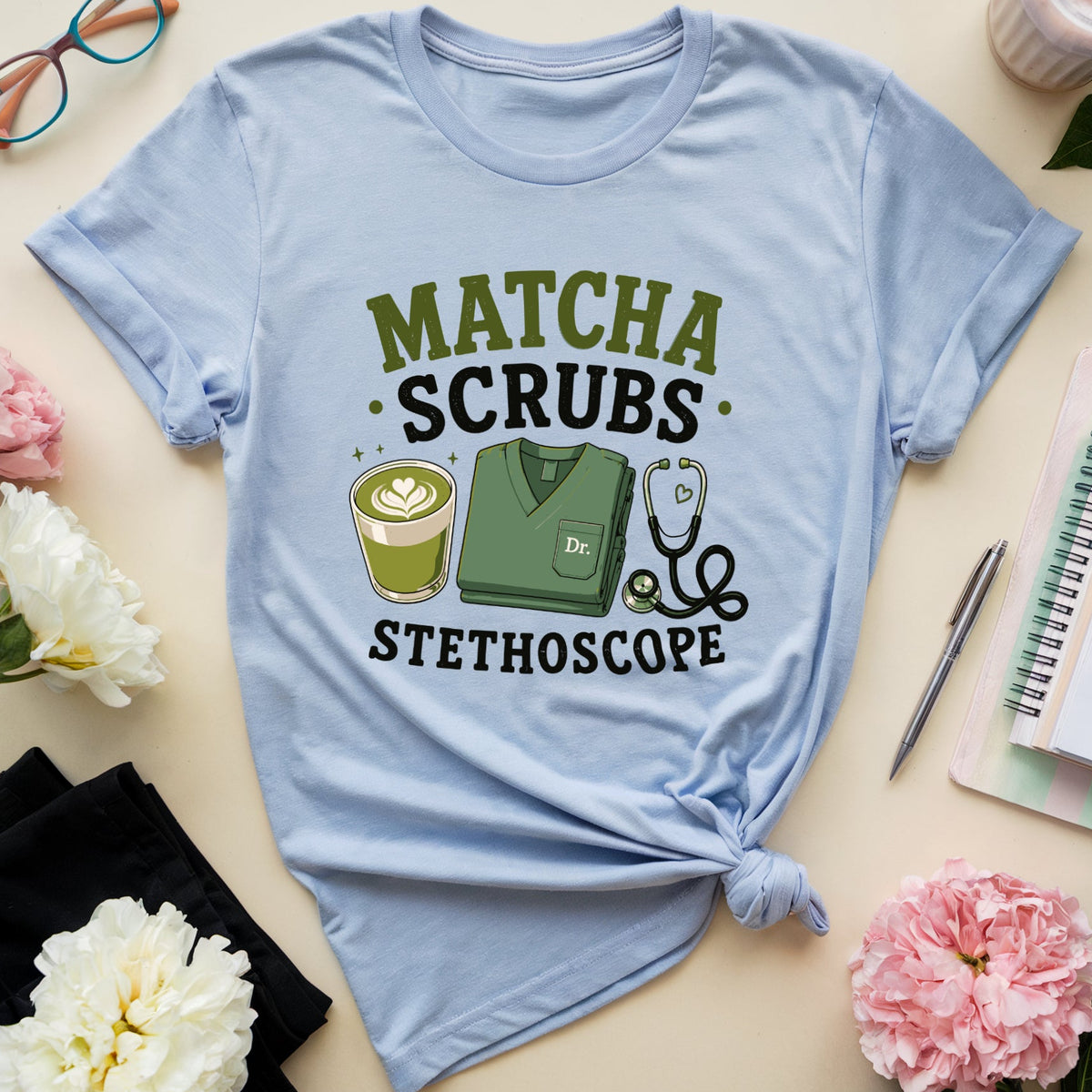 A light blue graphic t-shirt with rolled sleeves, featuring a 'MATCHA SCRUBS STETHOSCOPE' design with a coffee cup, scrub top, and stethoscope. The t-shirt is styled in a flat lay with flowers, glasses, a notebook, and a cup of iced coffee on a beige background.