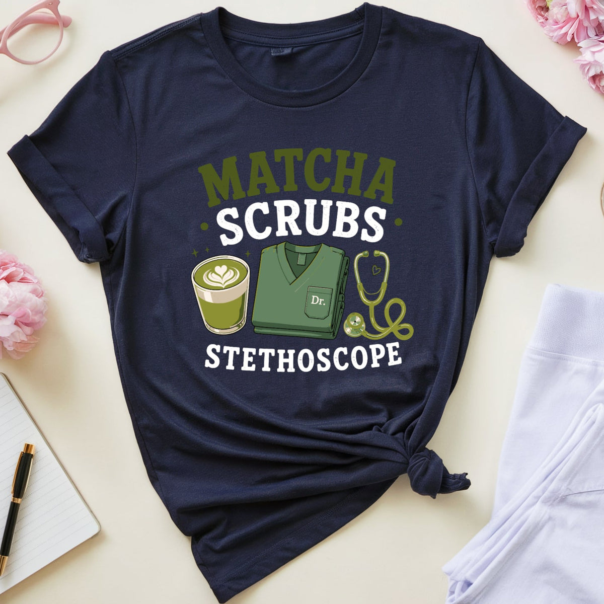 A navy blue graphic t-shirt styled in a flat lay with pink flowers, glasses, a notebook, a cup of tea, and folded white scrubs. The t-shirt features a 'MATCHA SCRUBS STETHOSCOPE' graphic with a stethoscope, book, and coffee cup illustration.
