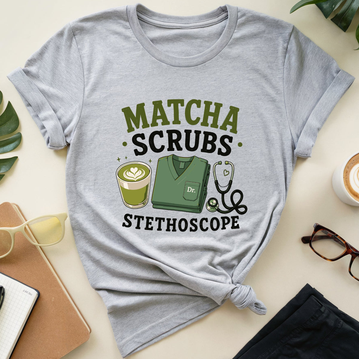 A heather gray graphic t-shirt styled in a flat lay with a knotted hem, featuring the text 'MATCHA SCRUBS STETHOSCOPE' and medical-themed illustrations. The shirt is surrounded by lifestyle props including a notebook, sunglasses, a cup of latte, glasses, and a plant on a light beige background.